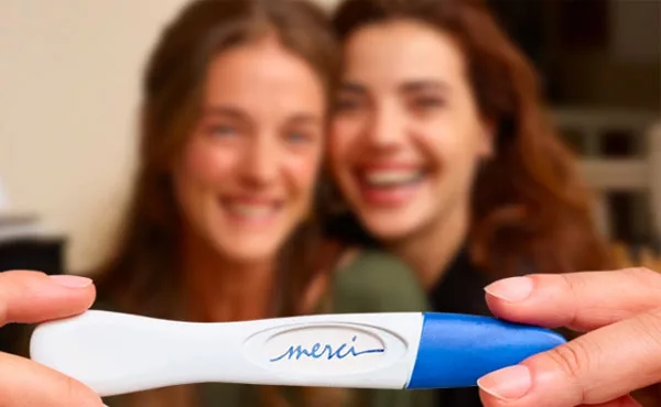 deux femmes sourient en arrière plan et montrent un test de grossesse sur lequel est écrit "merci"
